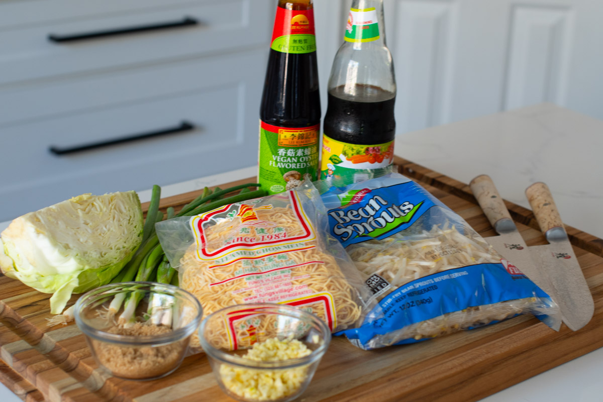 Fresh ingredients for Vegetarian Cambodian Chow Mein: cabbage, green onions, steamed chow mein noodles, bean sprouts, vegan oyster sauce, soy sauce, brown sugar, and minced garlic displayed on a wooden cutting board