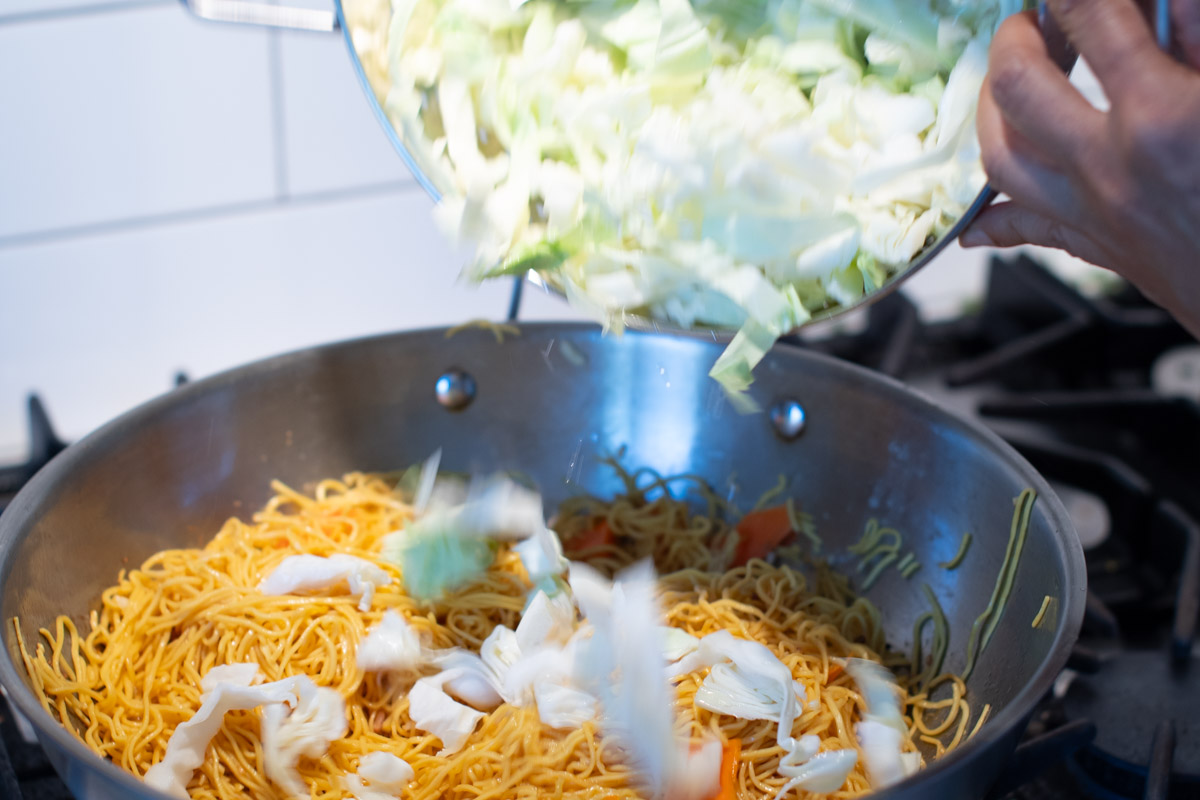 Mixing fresh vegetables into noodles during Cambodian stir-fry preparation.